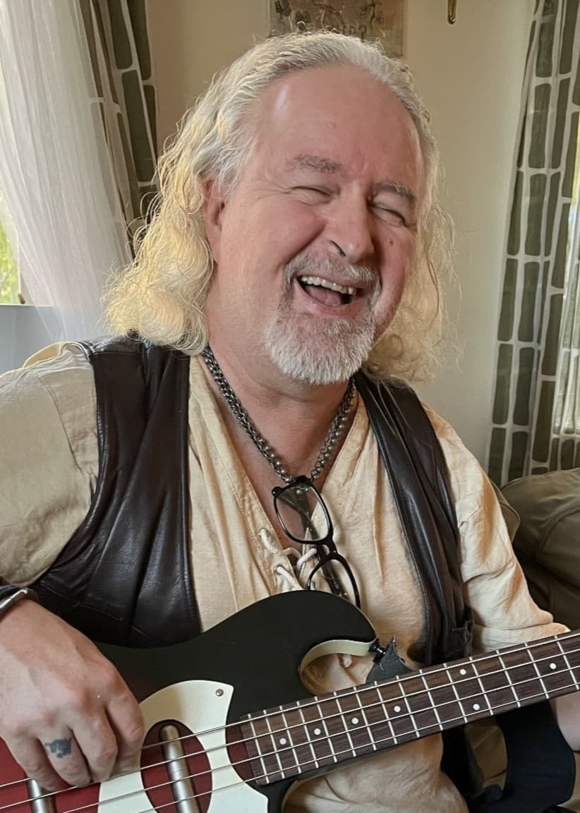 Stefan Pettersson spelar bas i ett vardagsrum med grönskande träd utanför fönstret. Rummet har ljusa gardiner, gröna kuddar och en avslappnad inredning.