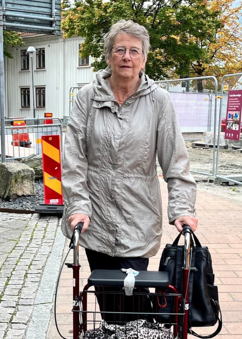 Britt Inger Eriksson går med en rullator på en trottoar intill ett pågående vägarbete med avspärrningar och byggstaket i bakgrunden.