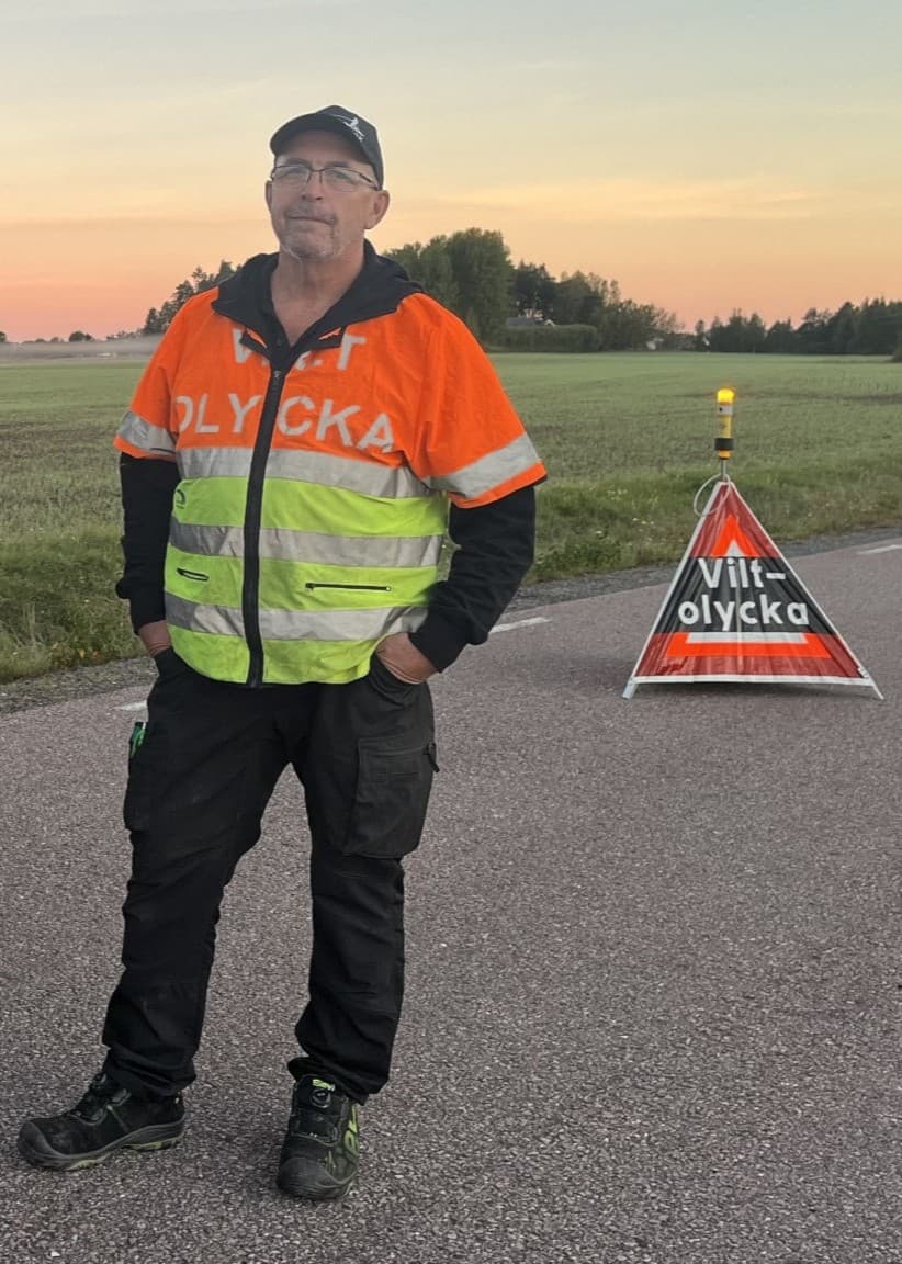 Eftersöksjägaren, Ingvar, står vid en svart bil med jaktutrustning på en landsväg. Han bär en reflexväst och bakom honom syns en varningsskylt samt öppet landskap i solnedgång.