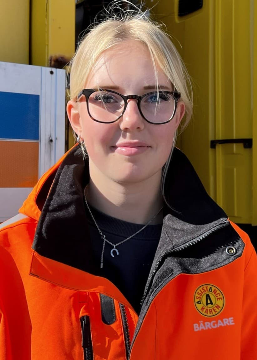 Alice Pettersson, klädd i en orange arbetsjacka, står framför en gul lastbil med blå och orange dekaler.