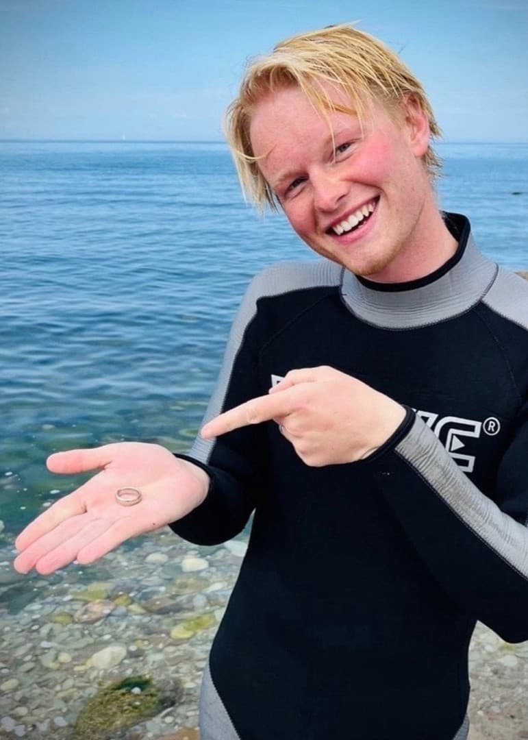 Elias Wärlinge står vid en strand på Öland i våtdräkt och pekar på ett litet föremål i sin hand, som tros vara hittat med en metalldetektor. En närbild av detektorn visas bredvid.