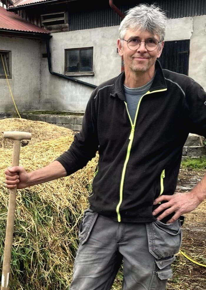 Jesper Sandström står utomhus vid en gård, klädd i arbetskläder och håller i en grep. Bakgrunden visar en hög med grödor och äldre ladugårdsbyggnader med slitna fasader.