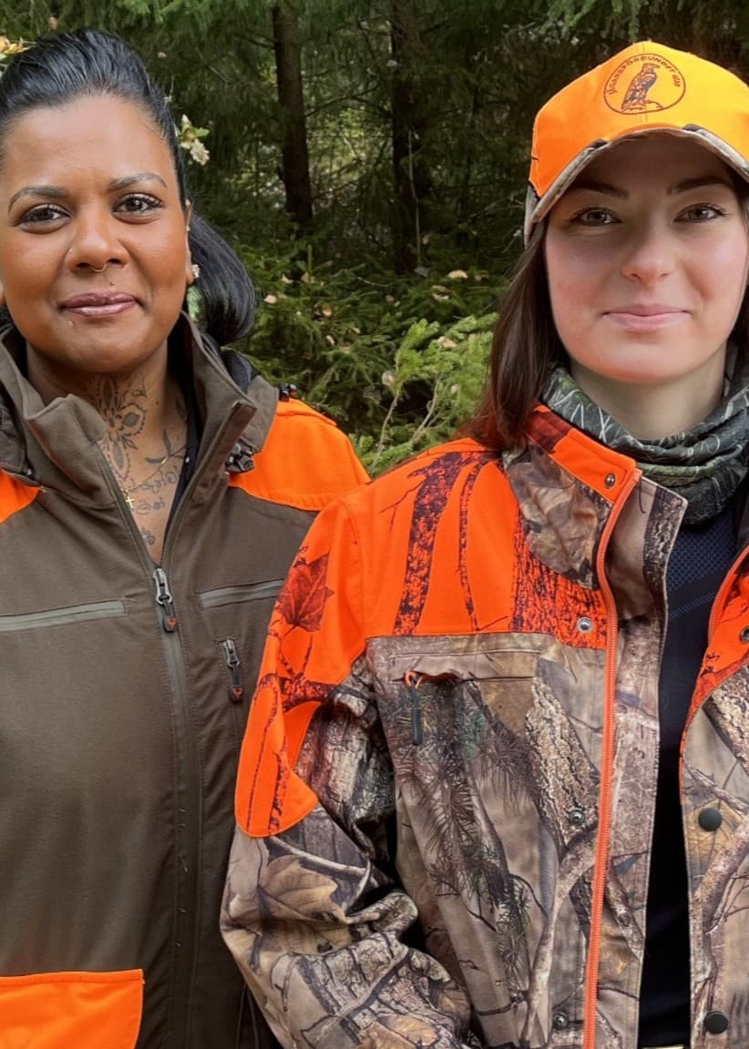 Två personer, Filippa och Nimali, iklädda jaktkläder med orange detaljer står utomhus framför grönskande skogsmiljö. Bilden framhäver jakt som naturaktivitet och deras utrustning.