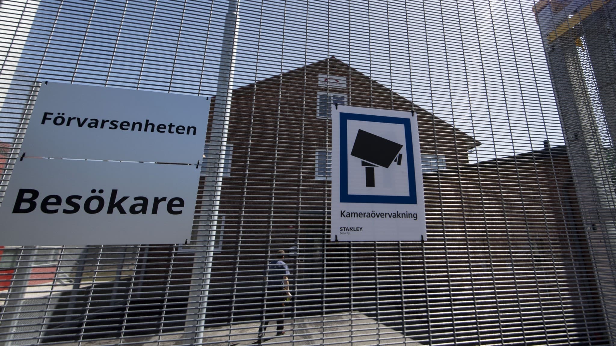 Migrationsverkets förvar i Ljungbyhed, omgärdat av högt stängsel med skyltar om övervakning. Byggnaden i bakgrunden används för att inhysa förvarstagna personer.