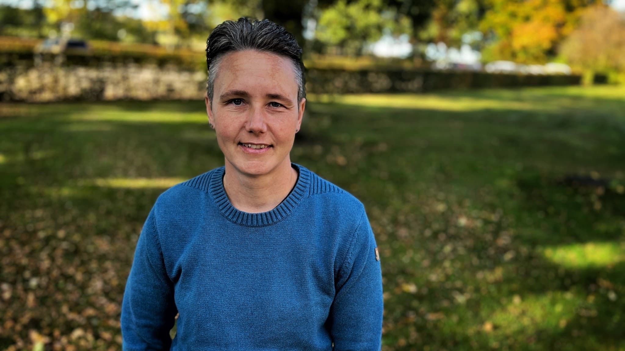 Elin Schwartz står utomhus i en park med höstlöv på marken och träd i bakgrunden. Personen har på sig en blå stickad tröja. 