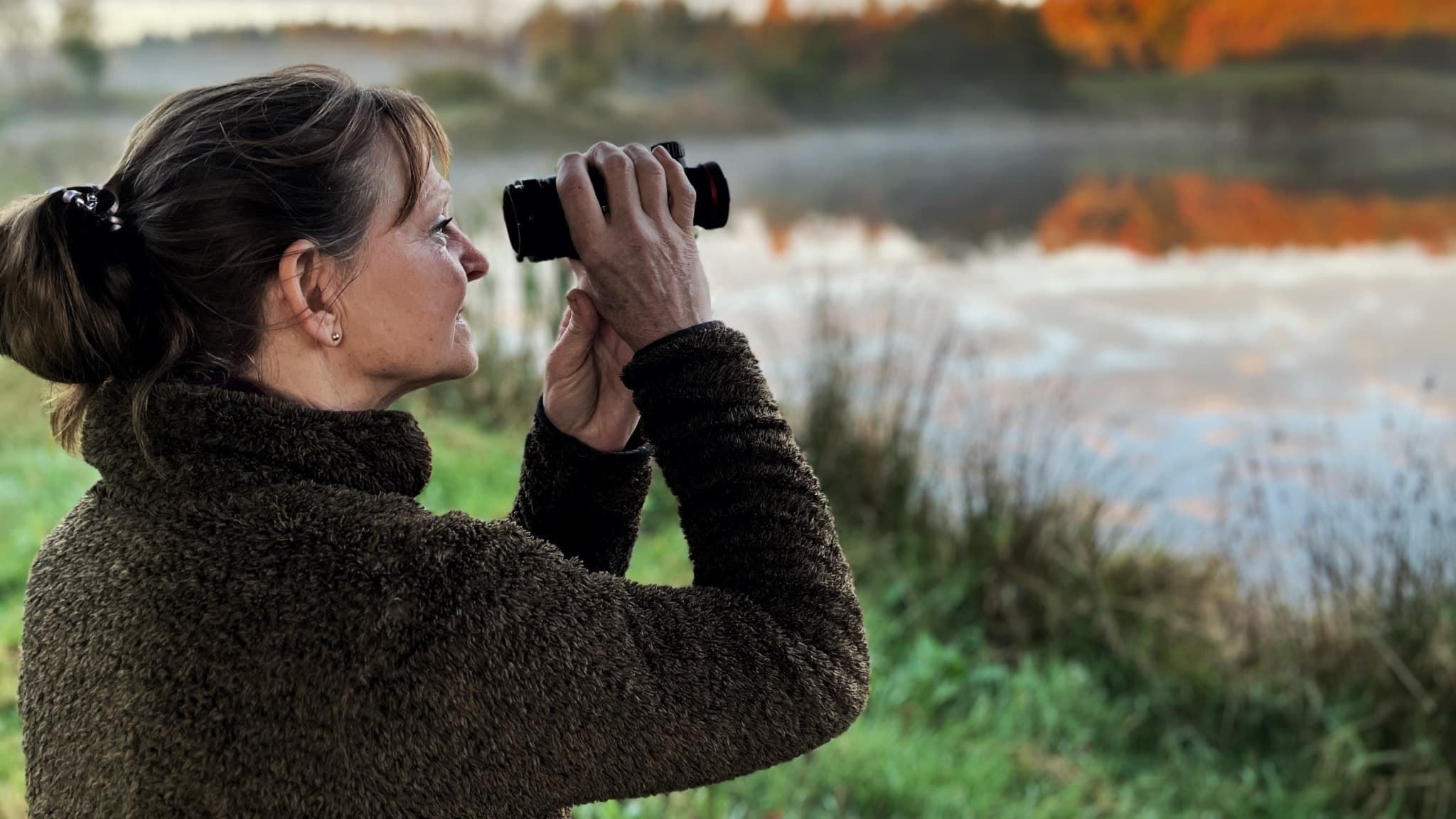 Anette Holst mörkt hår och mörk tröja står vid en sjö och tittar genom en kikare. Sjön omges av grönska och höstfärgade träd, med dimma som ligger över vattenytan.