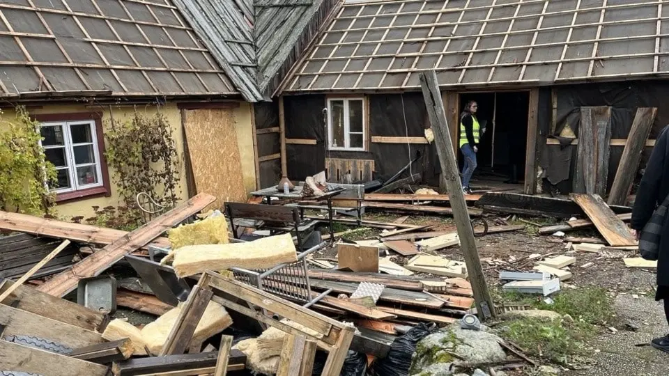 En äldre byggnad med träfasad har kollapsat delvis, med bråte och plankor utspridda på marken. En person i varselkläder syns vid en dörröppning.