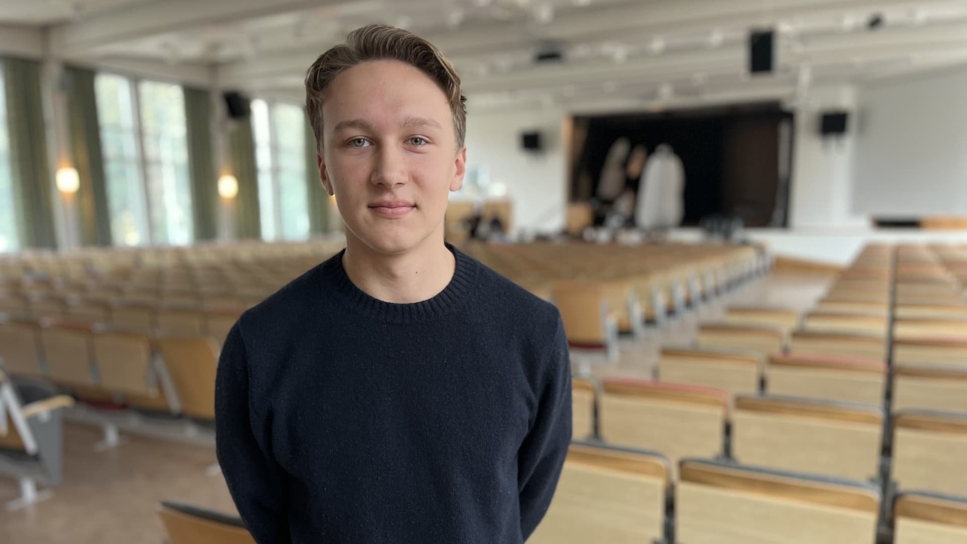 August Linell från elevkåren står i en aula med tomma stolsrader och en scen i bakgrunden på Söderportgymnasiet, där skolfotografering organiseras.