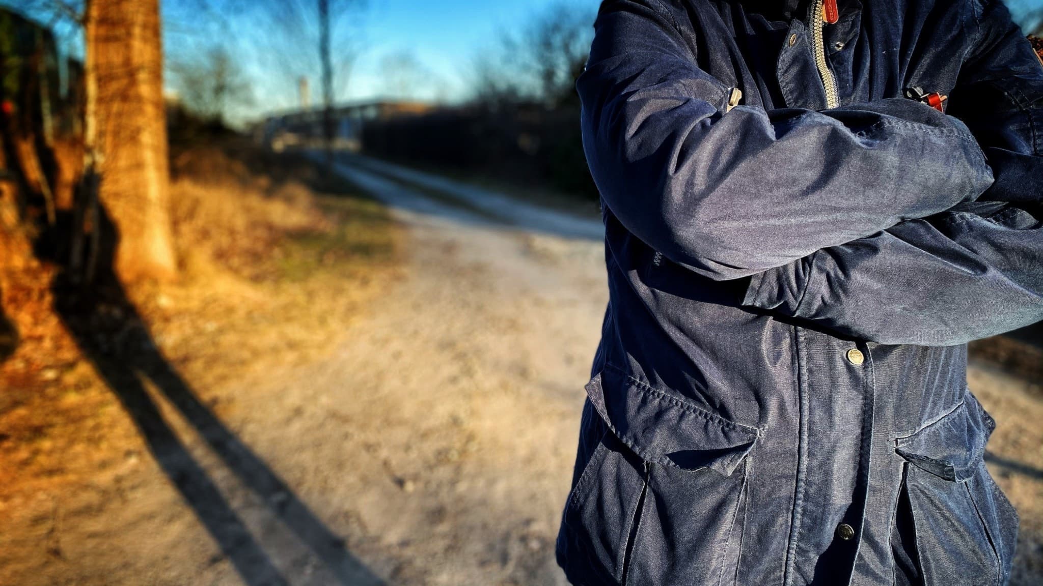 En kvinna i blå jacka står med korsade armar vid en grusväg i ett soligt men kyligt landskap.
