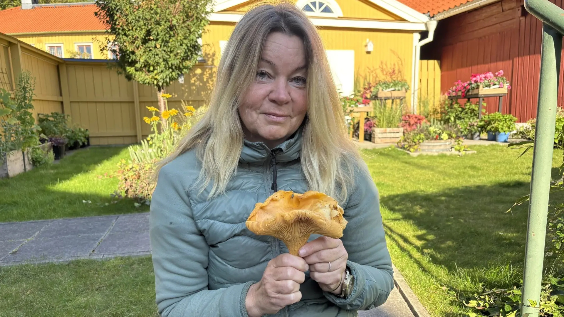 En person sitter utomhus i en trädgård med gulmålade hus i bakgrunden och håller upp en stor svamp i händerna, omgiven av grönska och höstväder.