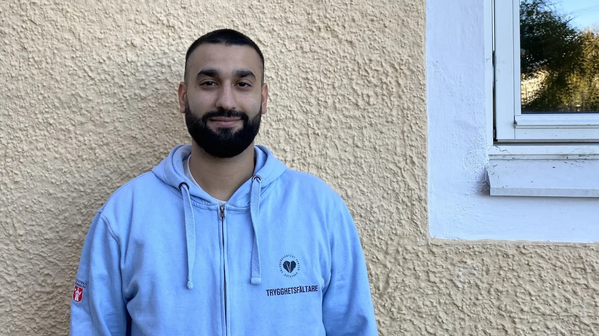 En person klädd i en ljusgrå hoodie står framför en beige fasad med två vita fönster på vardera sida. Bakgrunden visar en solig dag med grönska reflekterad i fönstren.