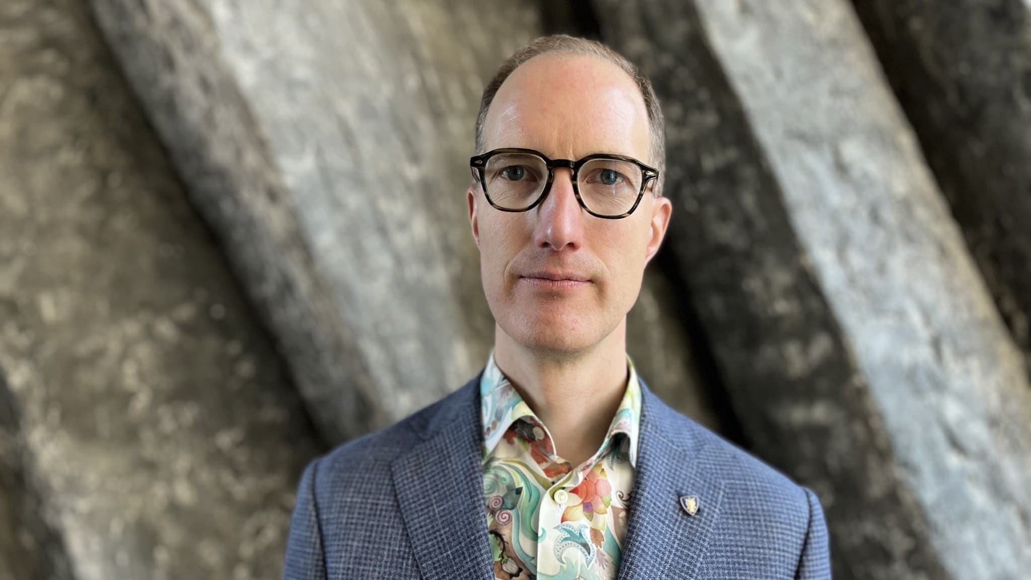 Liberalernas Jan Jönsson klädd i en blå kavaj med rutmönster och en färgglad skjorta står framför en bakgrund av lutande betong.