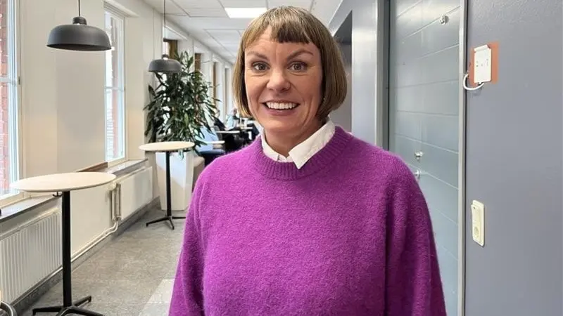 Maja Österlund, projektledare, står i en modern kontorsmiljö med vita väggar, stora fönster och grå dörrar. Hon bär en lila tröja och vit skjorta. Miljön är ljus och minimalistisk.