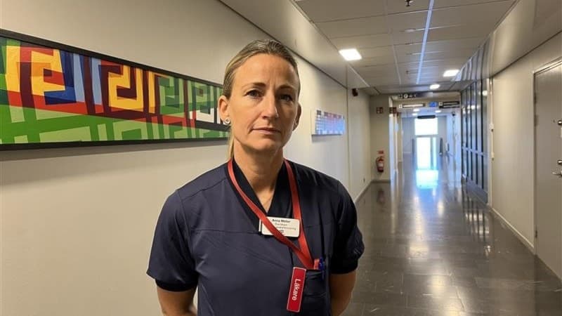 En person i mörkblå arbetskläder står i en sjukhuskorridor med en färgglad tavla på väggen. Personen har ett ID-kort runt halsen och korridoren sträcker sig mot en glasdörr i bakgrunden.