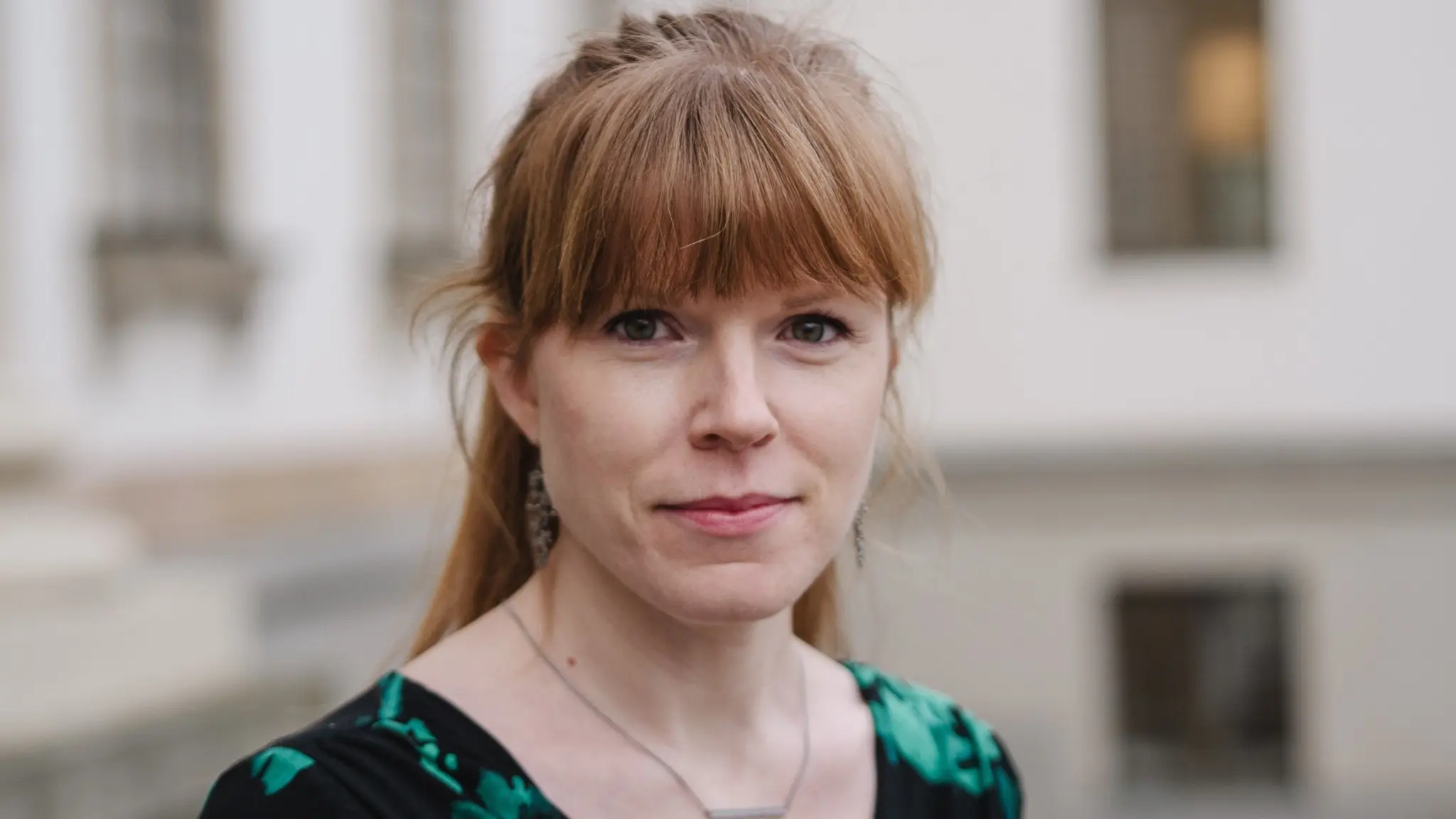 En kvinna med ljust hår och en grön mönstrad klänning står utomhus framför en byggnad med stora fönster och vita väggar, vilket kan indikera en officiell eller offentlig miljö.