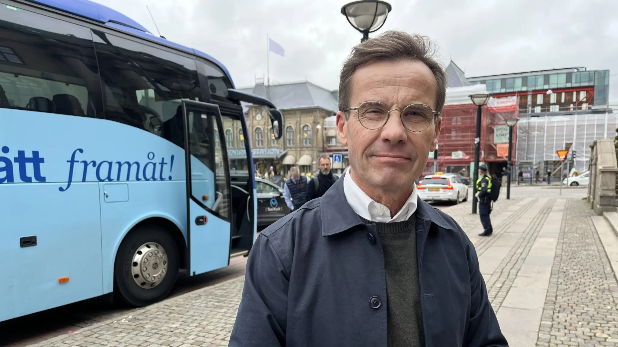 En person står framför en blå buss med texten 'Framåt' på sidan, på en trottoar i stadsmiljö. I bakgrunden syns byggnader, folk och en mulen himmel.