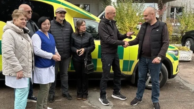 En grupp människor står framför en gulgrön ambulans som har donerats till ett sjukhus i norra Ukraina av Hönö Missionskyrka och Sionförsamlingen Rörö. Två personer skakar hand.