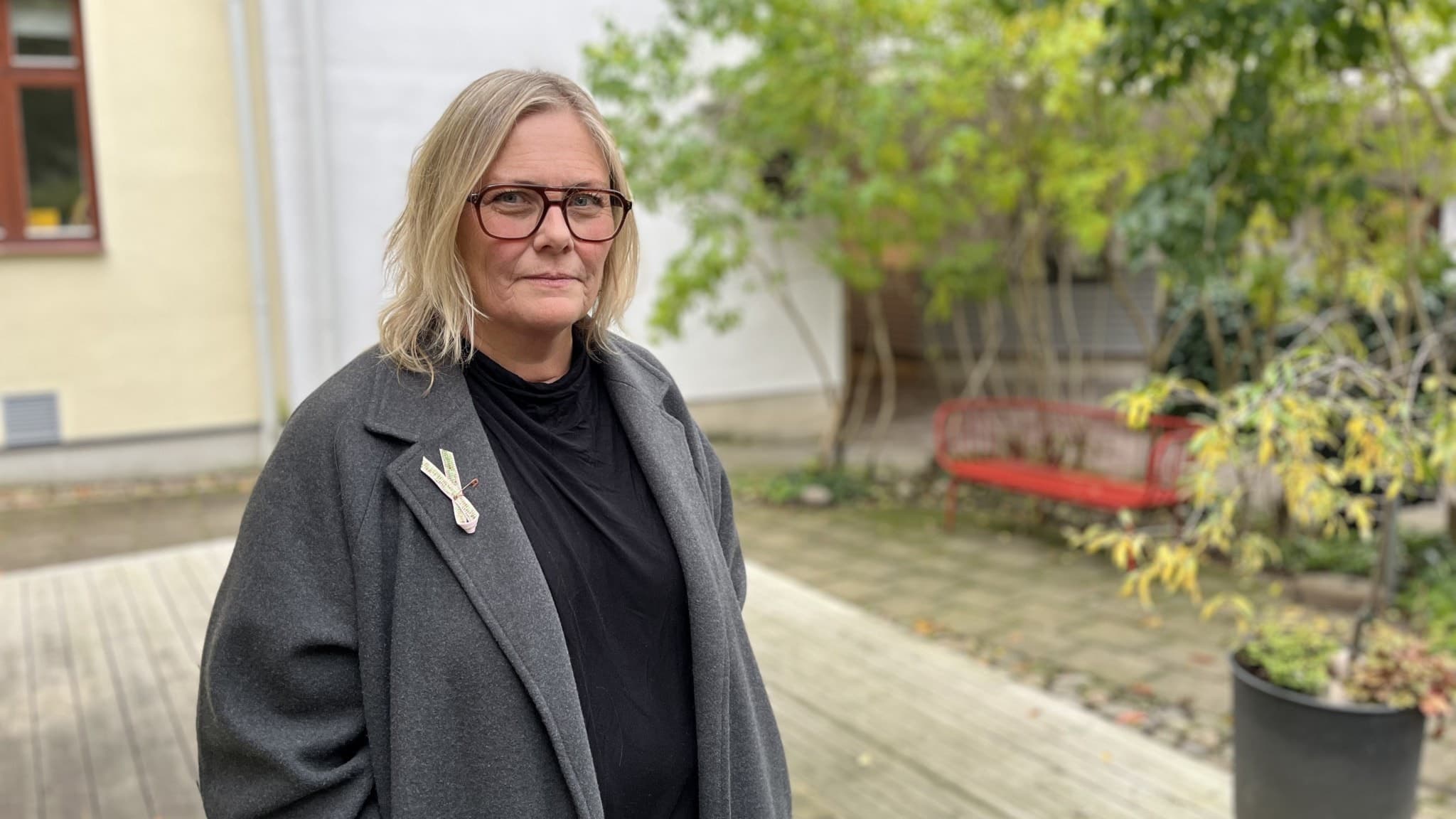 En person står utomhus på en träplattform, omgiven av grönskande träd och en byggnad med beige fasad och fönster i bakgrunden. Personen är klädd i en mörk kappa och svart tröja.
