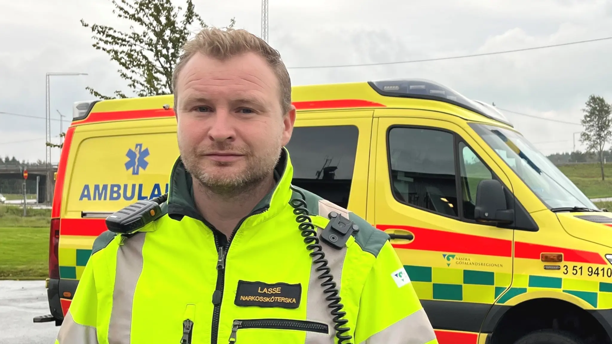 En man i gul varseljacka framför en ambulans vid ett grönområde.
