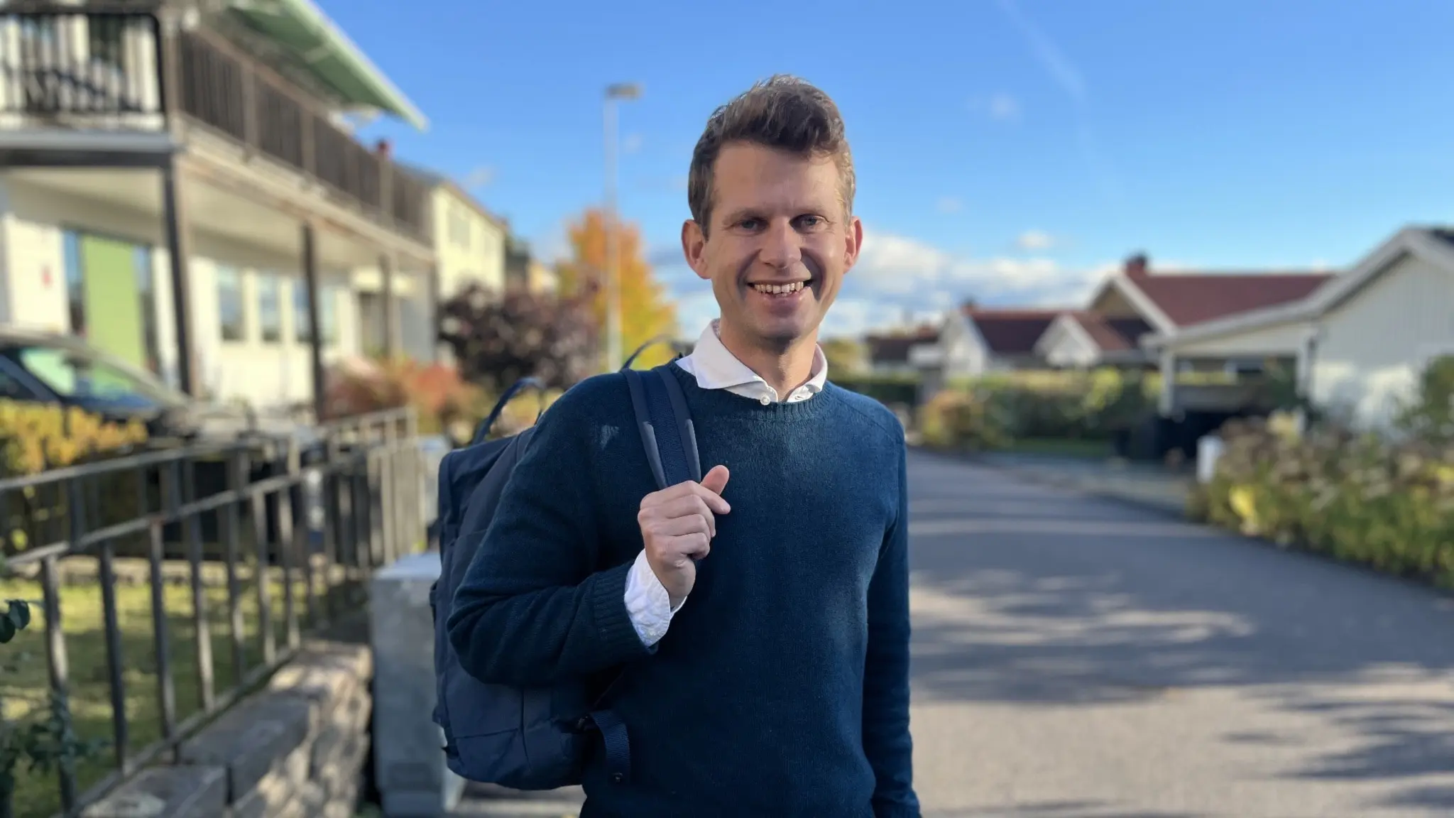 Axel Darvik (L) står utomhus på en villagata med ryggsäck och höstklädd i blå tröja. Hus med trädgårdar syns i bakgrunden under en klarblå himmel.