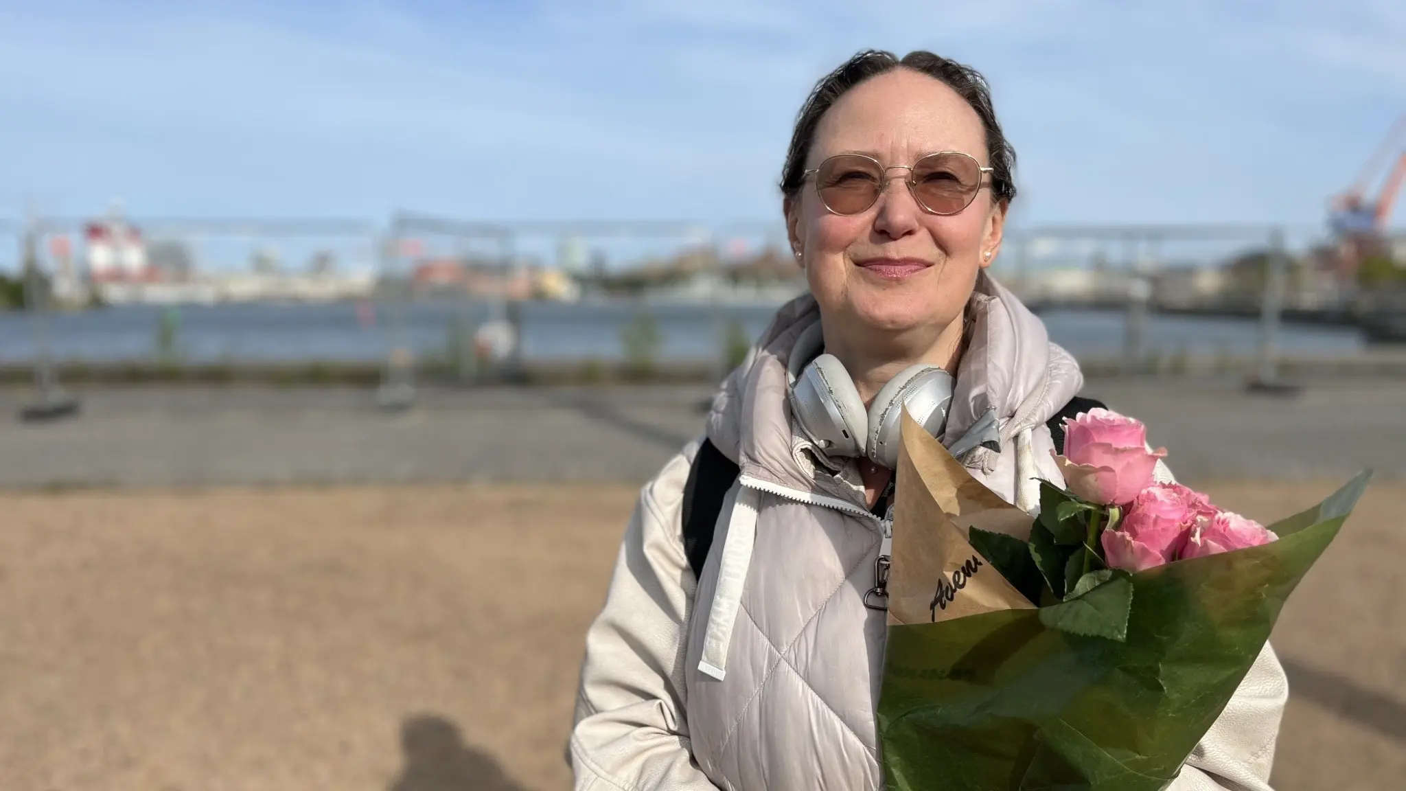 En kvinna med solglasögon och hörlurar om halsen står på kajen med ett fång rosor i famnen