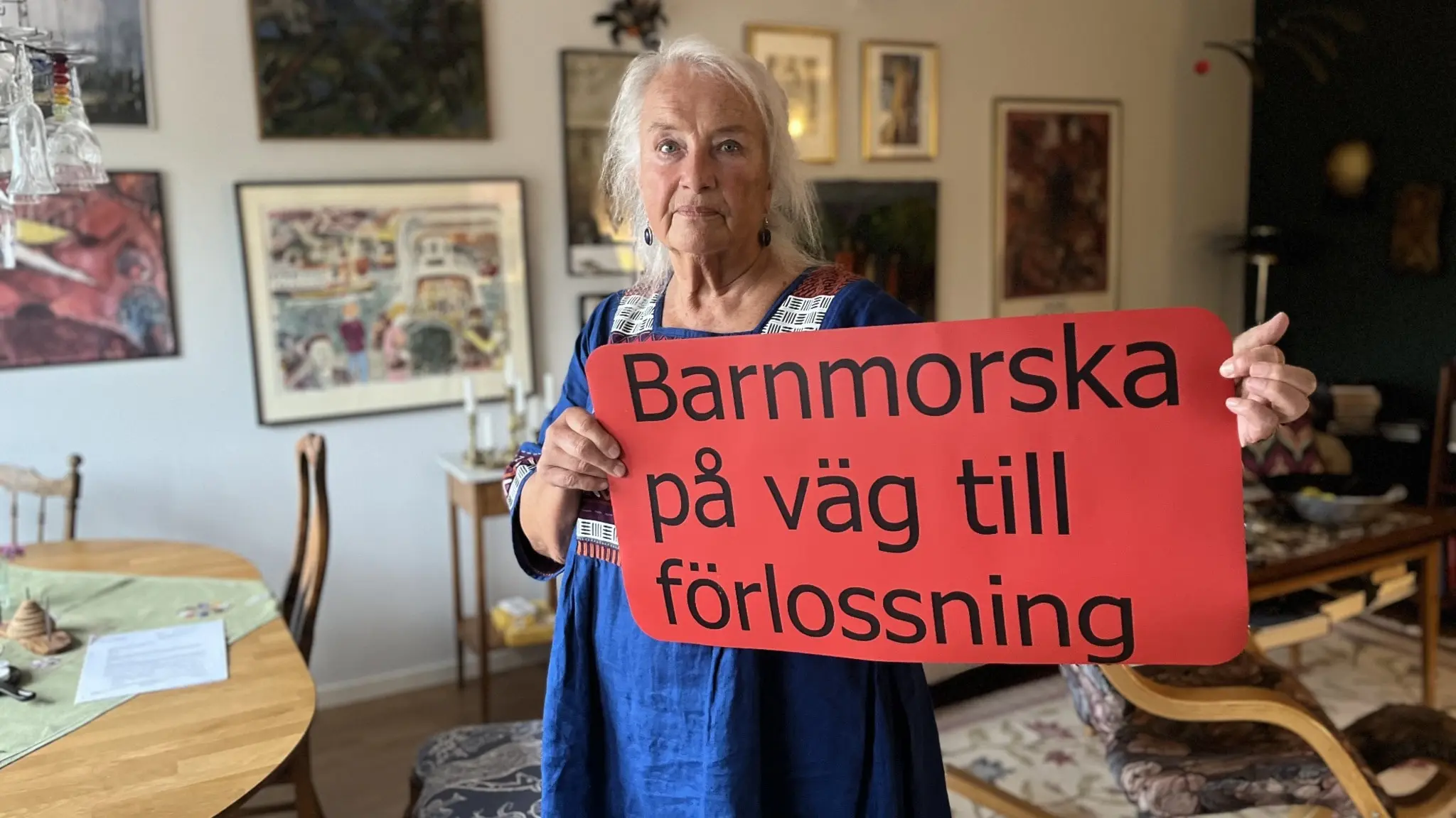 En person håller upp en röd skylt med texten 'Barnmorska på väg till förlossning' i ett hemtrevligt vardagsrum med konst på väggarna och möbler i trä.