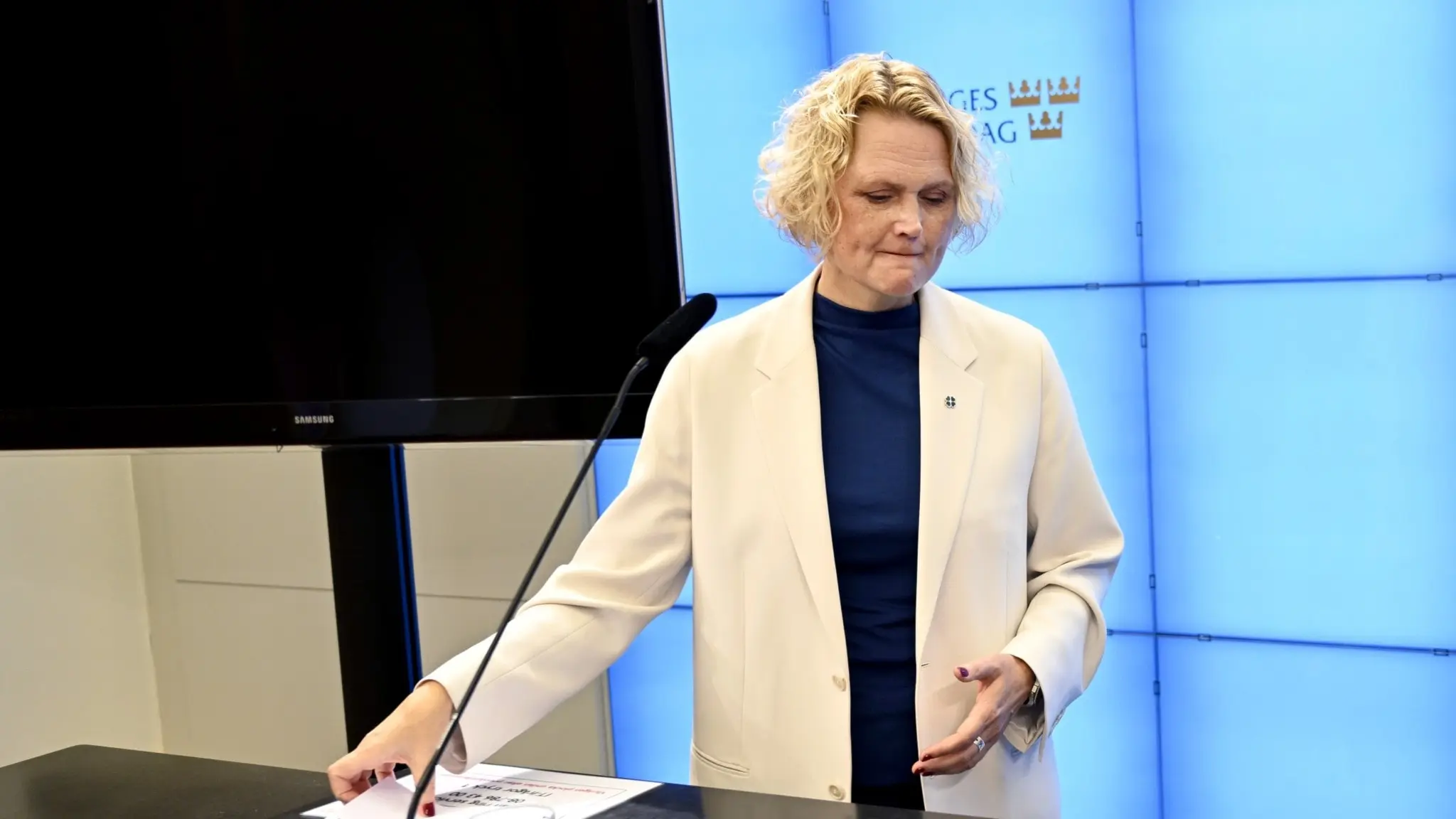 Centerpartiets partiledare Anna-Karin Hatt står vid ett podium under en pressträff i Stockholm. Hon har på sig en vit kavaj och blå topp, med papper och ett glas vatten framför sig.