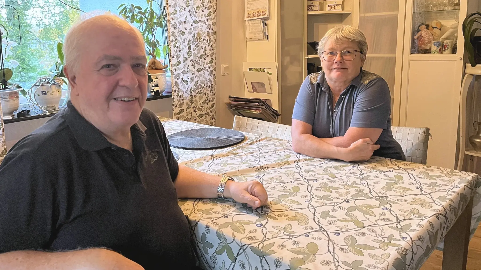 Jan-Erik och Anne-Marie Johannesson sitter vid ett köksbord med blommigt dukmönster i ett hem i Lerum. Fönstret i bakgrunden visar gröna växter och gardiner med blommigt mönster.