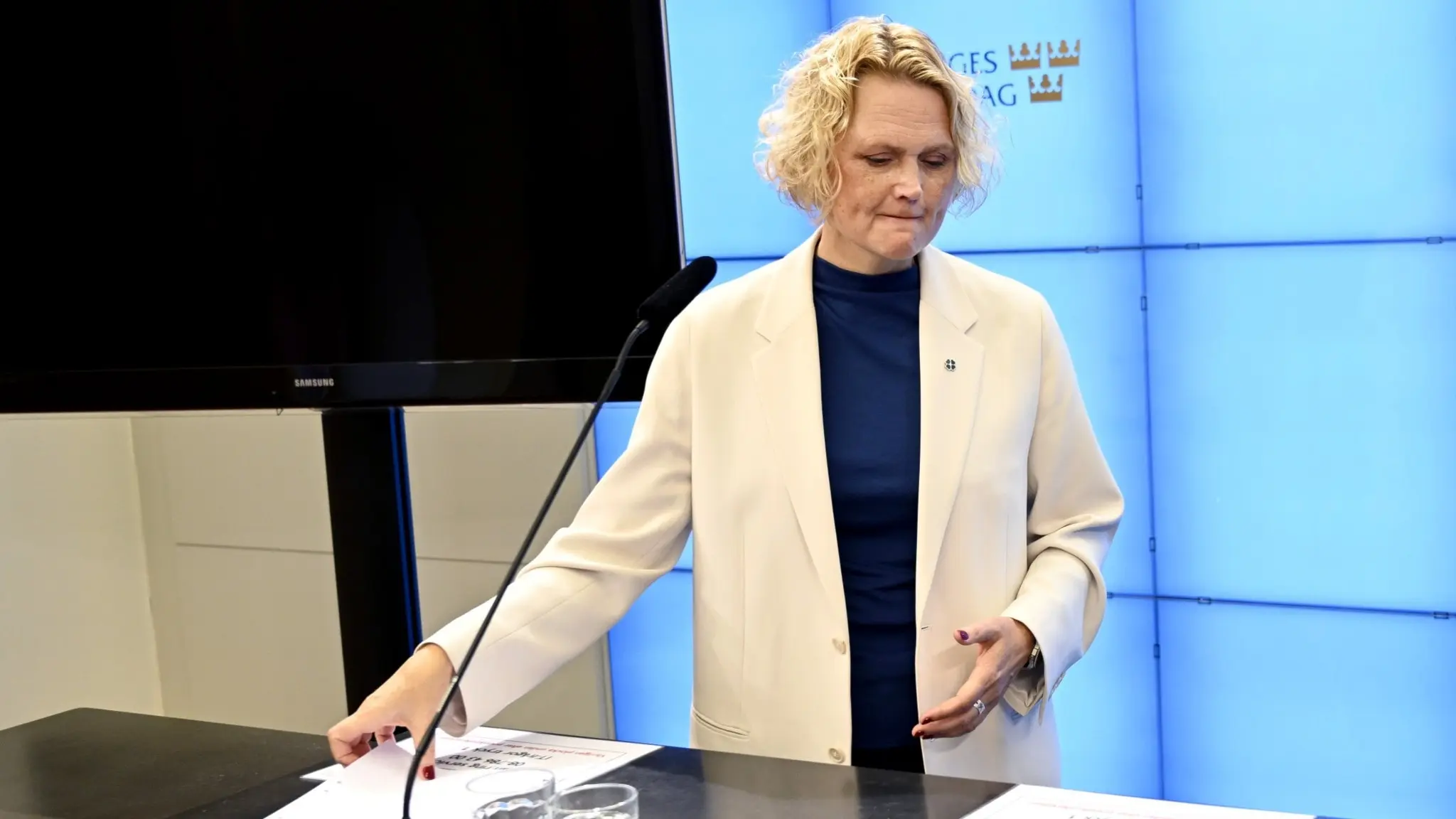 Centerpartiets partiledare Anna-Karin Hatt står vid ett podium under en pressträff i Stockholm, iklädd vit kavaj och blå tröja, med en blå digital bakgrund och papper på bordet.