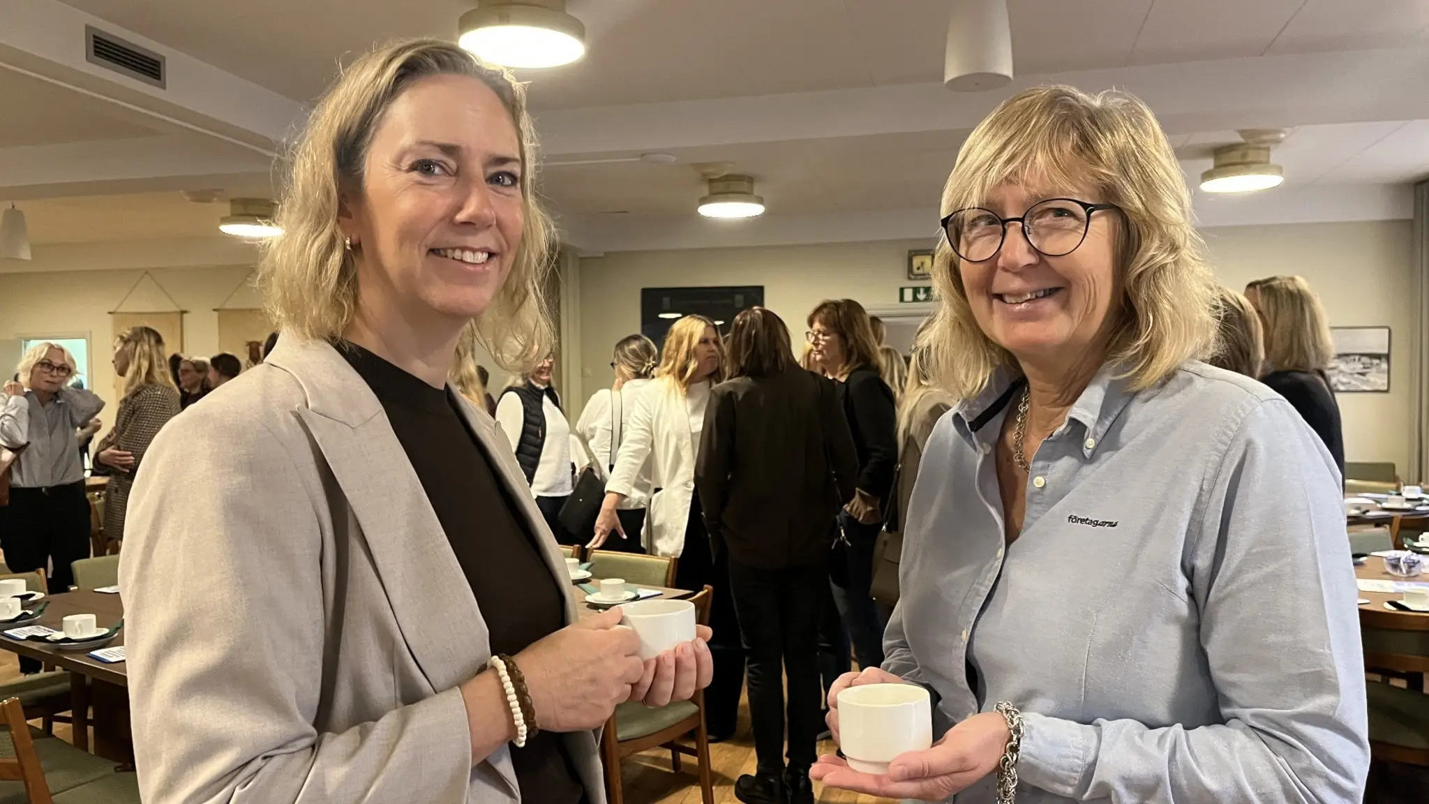 Elisabeth Dahl och Tina Ivarsson står i förgrunden och samtalar med kaffekoppar i händerna under Företagarnas kvinnofrukost. Bakgrunden visar flera deltagare i en konferensmiljö.