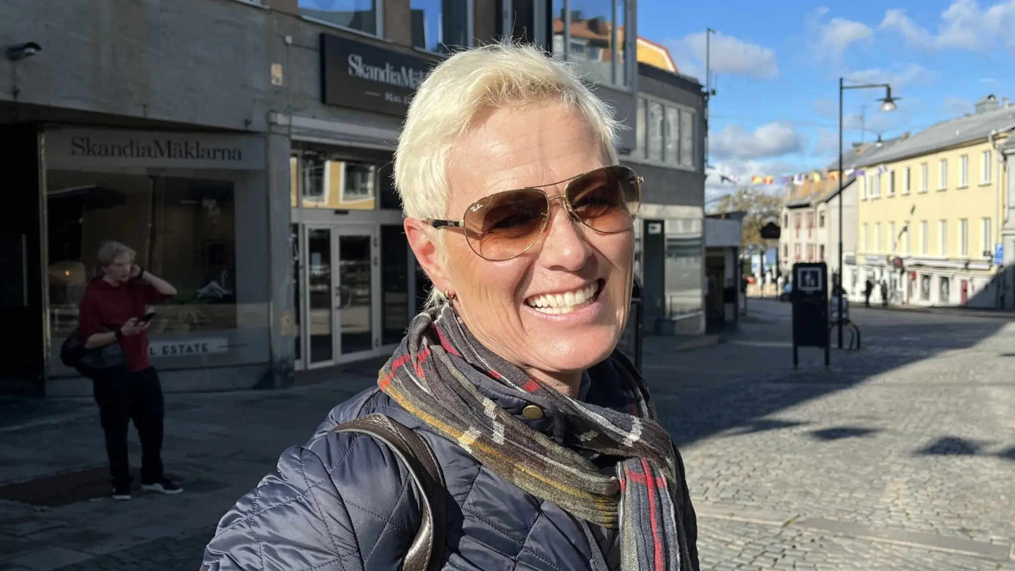 Sara Forell står utomhus på ett torg med blå himmel i bakgrunden. Hon bär mörk jacka och halsduk, omgiven av byggnader och öppet stadslandskap.