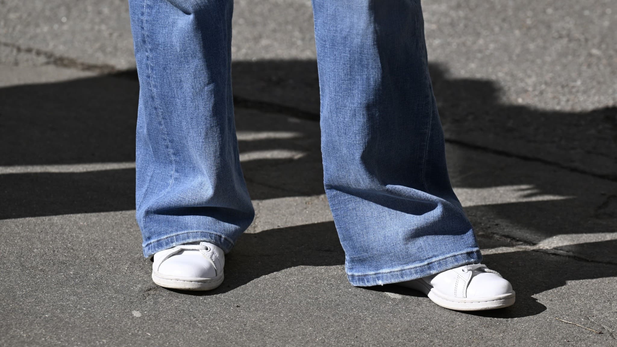 En person står utomhus på asfalt, klädd i utsvängda blå jeans och vita sneakers. Skuggan av personen syns på marken, vilket tyder på soligt väder.