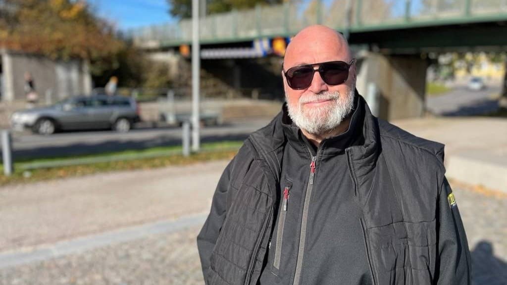 En person i mörk jacka står utomhus framför en bro med trafik i bakgrunden. Det är en solig dag med några moln på himlen. Höstfärger syns i träden runtomkring.