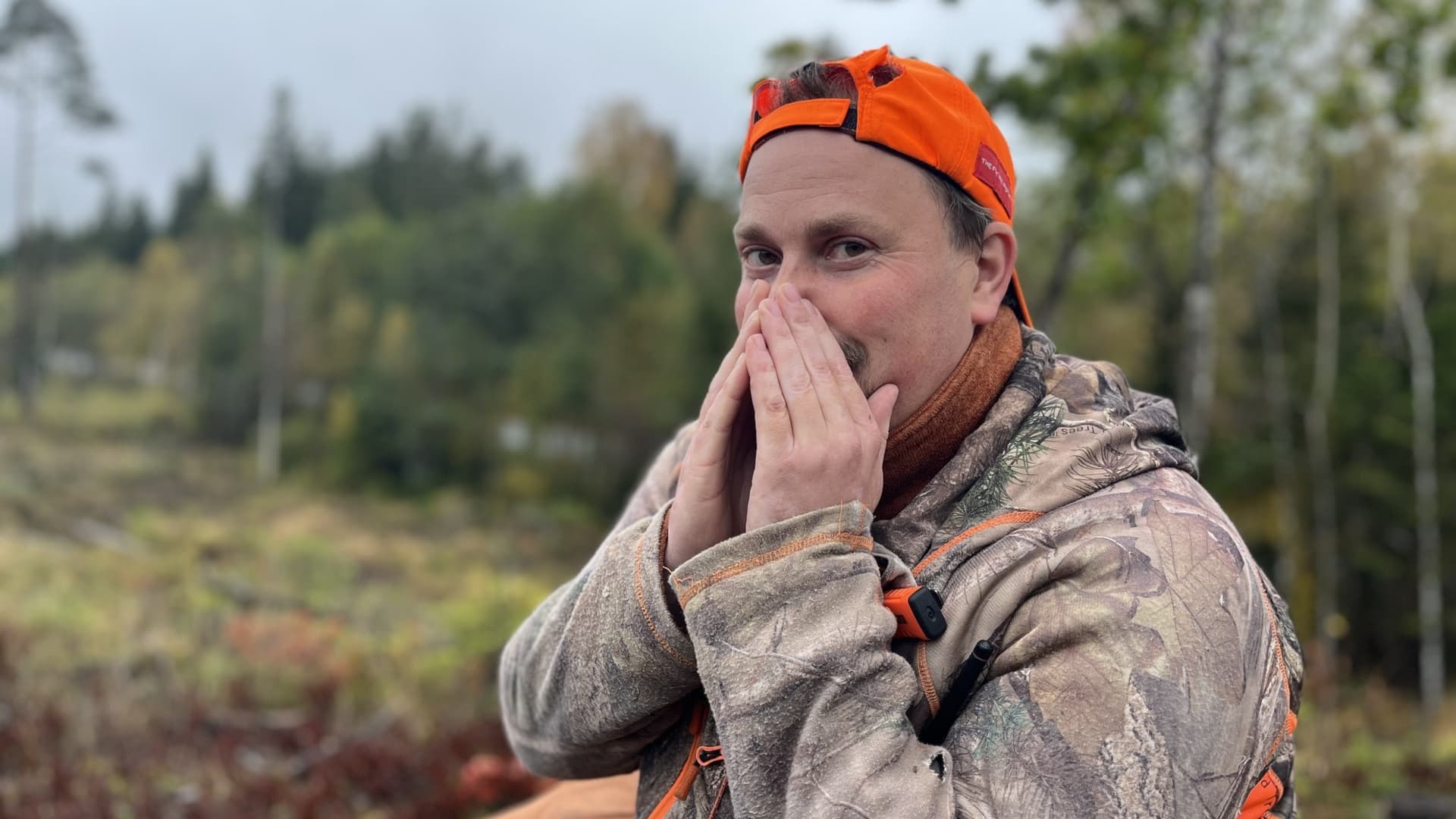 Gustaf sitter i jaktkläder och orange keps i skogsmiljö och använder ett knep för att locka älgar, omgiven av höstliga träd och en öppen glänta. Jaktknepet: Gustav kåtar upp älgarna