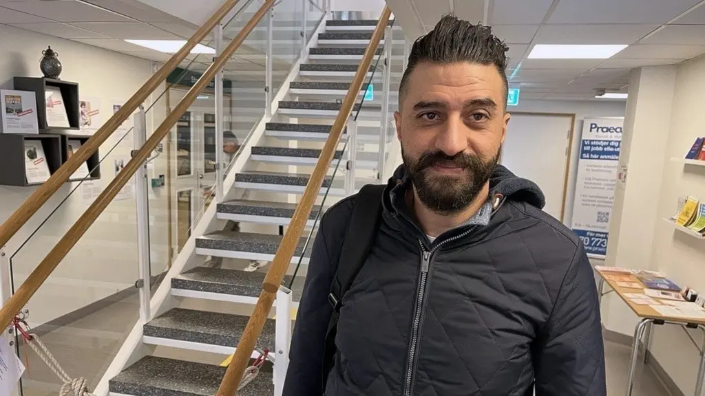 En man står inomhus framför en modern trappa med glasräcke. Lokalen har vita väggar, informationsmaterial på bord och hyllor samt skyltar vid entrén. Han bär en mörk jacka.