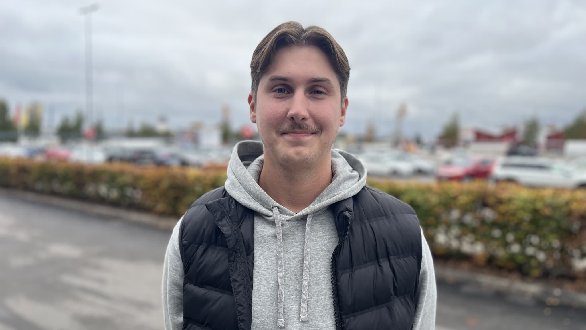 En person står utomhus på en parkeringsplats med bilar och buskar i bakgrunden. Personen bär en grå hoodie och en svart väst. Himlen är molnig och miljön har höstliga färger.