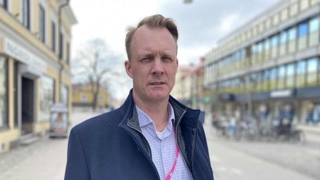Mikael Johansson står utomhus på en gata med byggnader i bakgrunden. Han bär en mörk kappa och skjorta med ett rosa band runt halsen som antyder tjänst eller uppdrag.