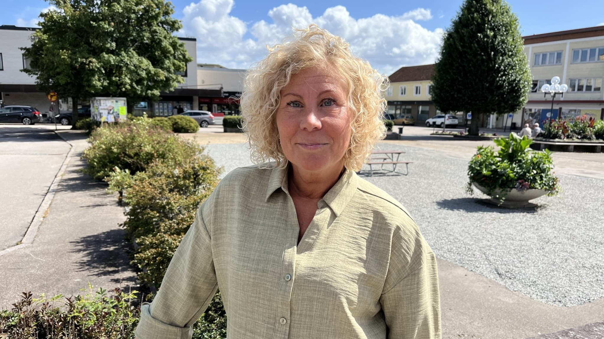 En kvinna med lockigt blont hår står utomhus på ett torg med grönskande buskar och träd i bakgrunden. Byggnader omger torget under en klarblå himmel med vita moln.