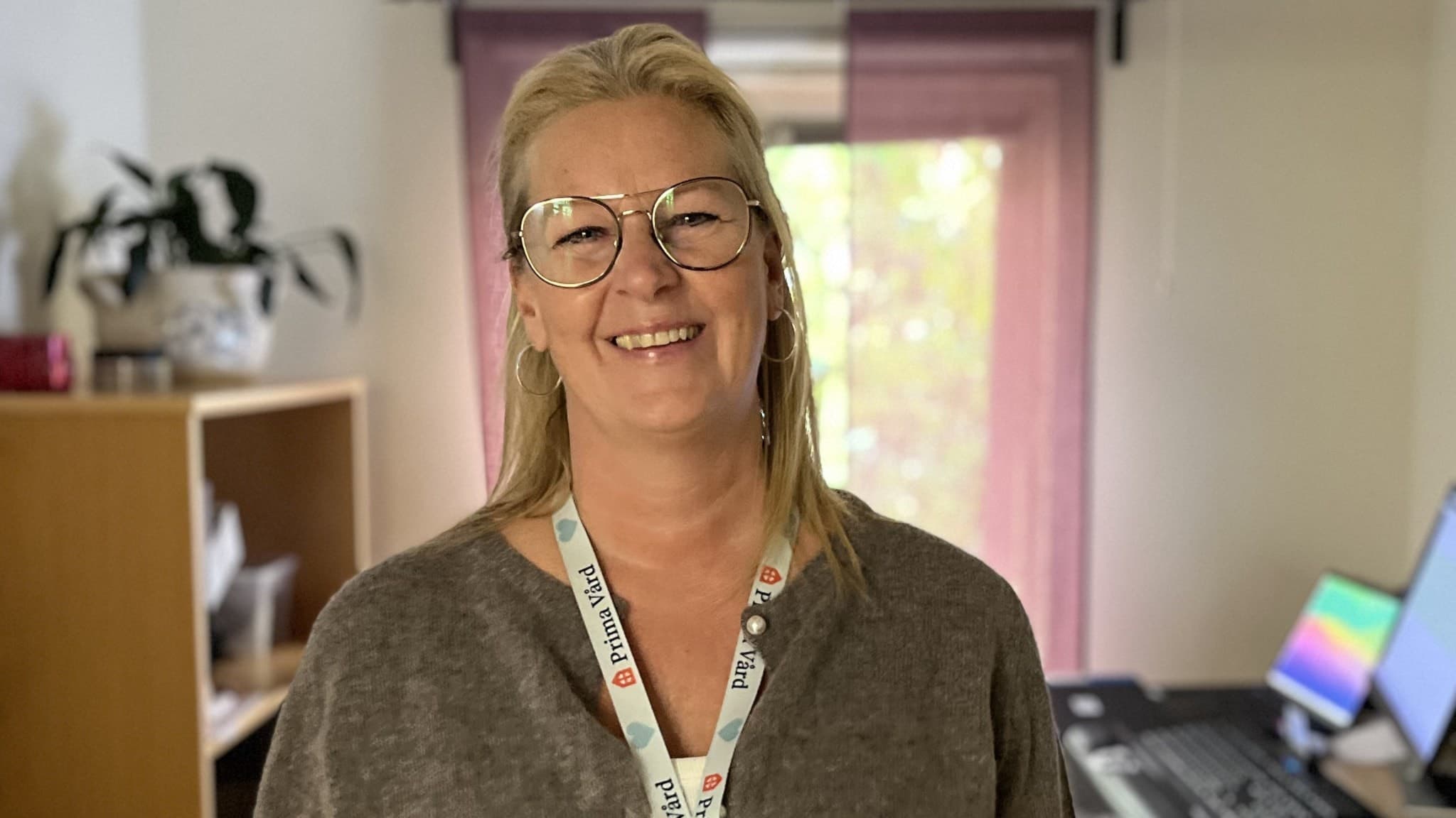 En person står i ett kontorsrum med hyllor fyllda med broschyrer till vänster och ett skrivbord med datorskärmar till höger. Rummet har ljusinsläpp från ett fönster med lila gardiner.