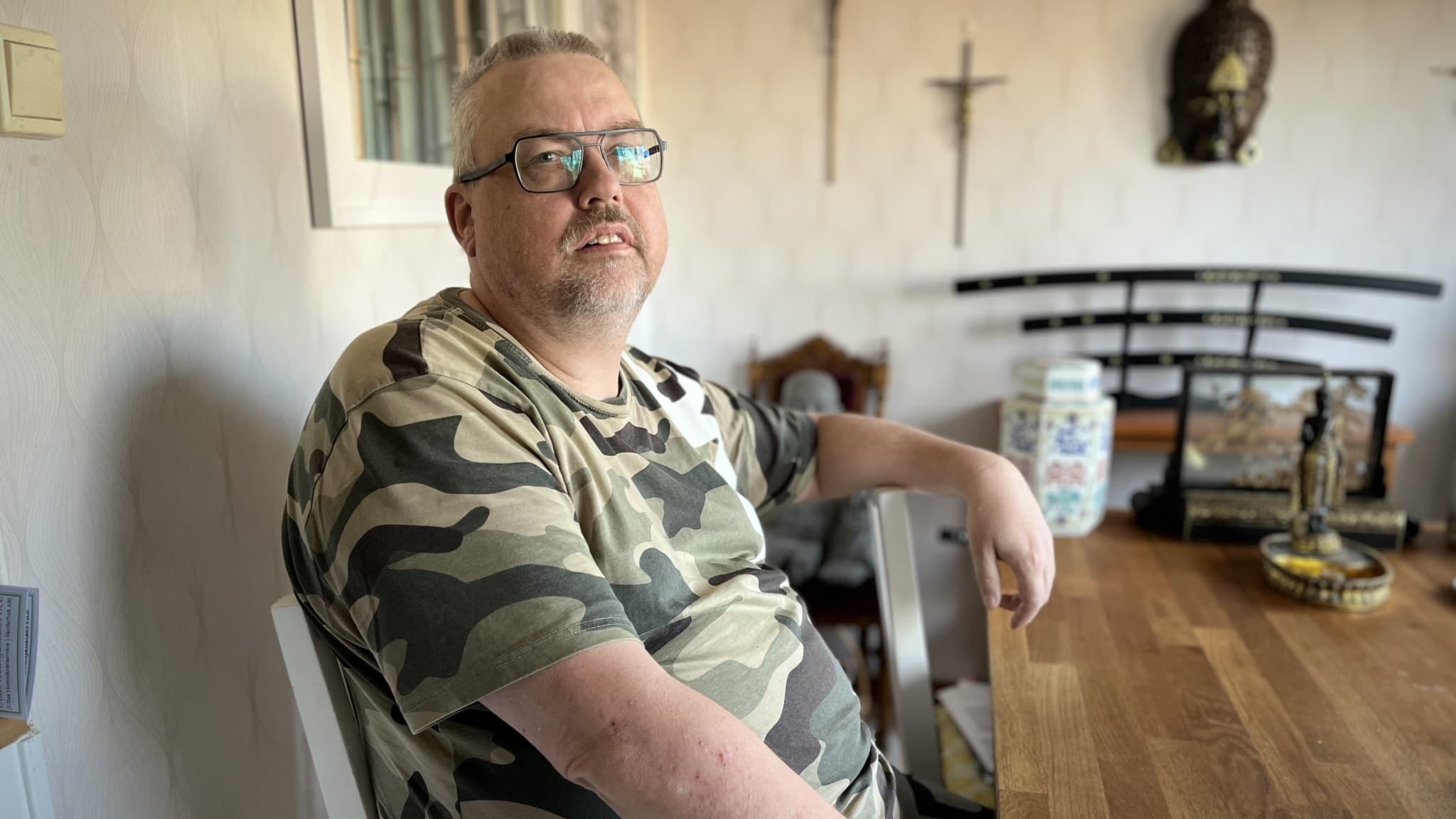 Per Lundberg sitter vid ett träbord i ett rum med religiösa dekorationer, inklusive kors och en staty. Han bär en kamouflagefärgad tröja och vilar sin arm på en stol.