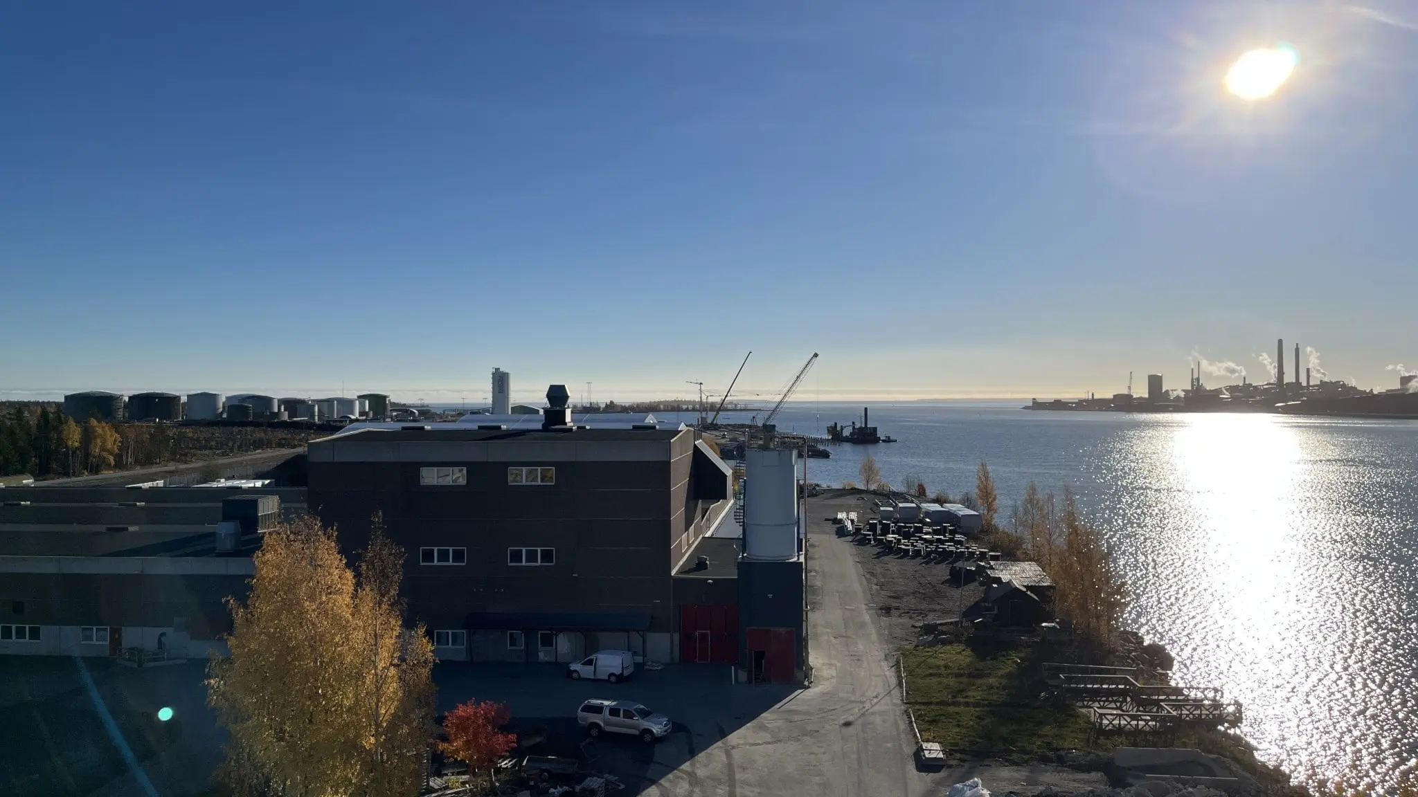 Solig vy över Skelleftehamns hamn med industribyggnader, kranar och båtar. Vattnet glittrar i solskenet och höstfärgade träd syns i förgrunden.