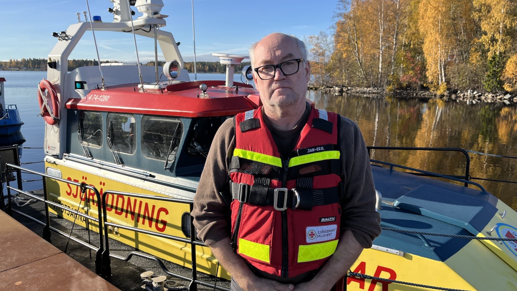 Jan Egil Hemfjell, skeppare på Sjöräddningssällskapet i Skellefteå, står framför en räddningsbåt vid en kaj, omgiven av höstfärgade träd och lugnt vatten.