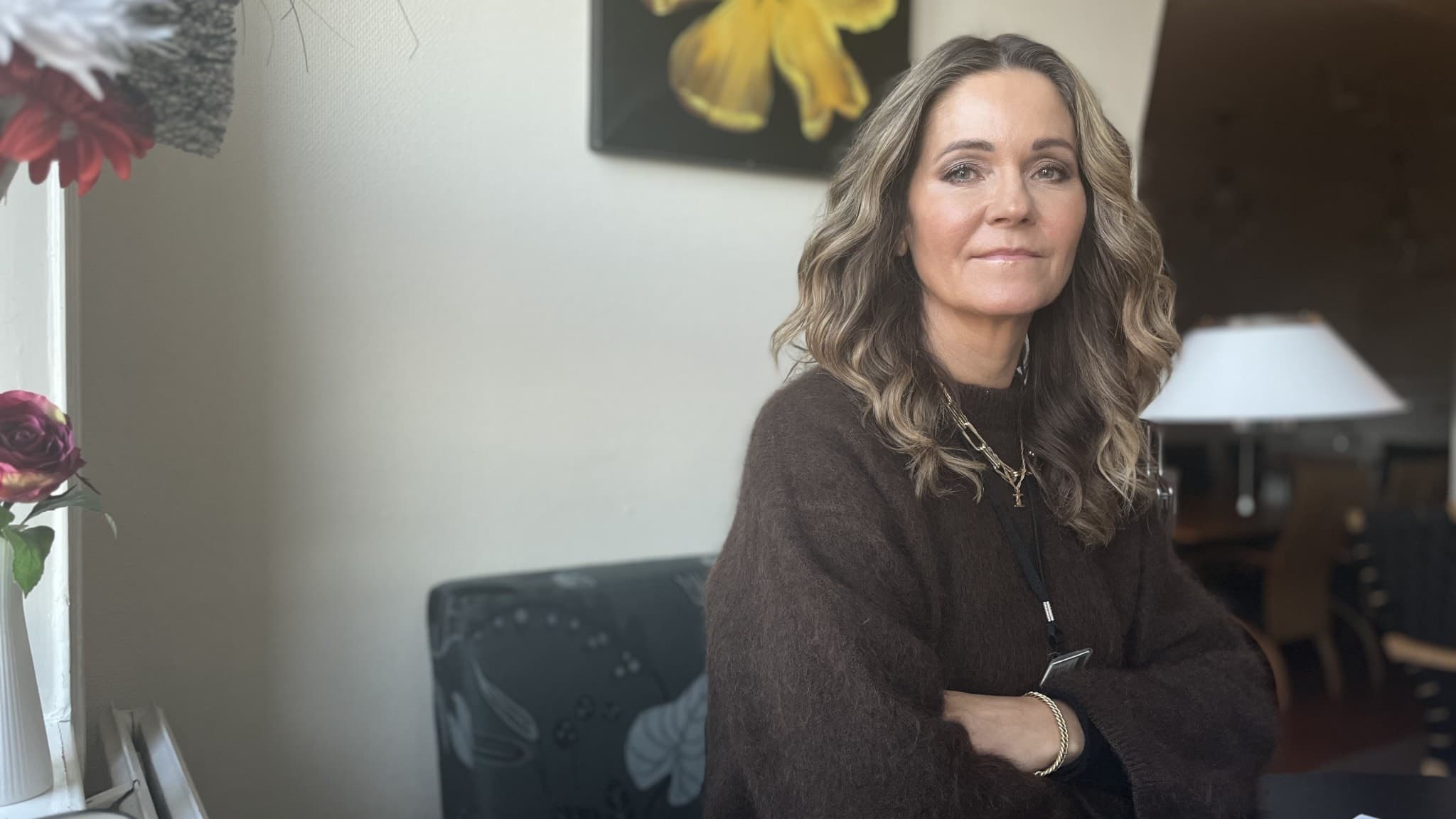 Pernilla Henriksson sitter vid ett bord i ett rum med en gul blomma på väggen och blommor i förgrunden. Hon bär en mörk tröja och ett halsband.