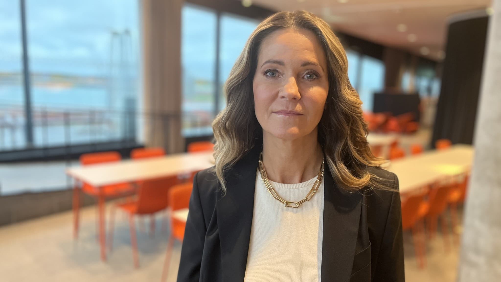 Pernilla Henriksson står inomhus i en miljö med stora fönster och röda stolar i bakgrunden. Hon bär en vit tröja och en svart kavaj, med vågigt hår och guldsmycken.