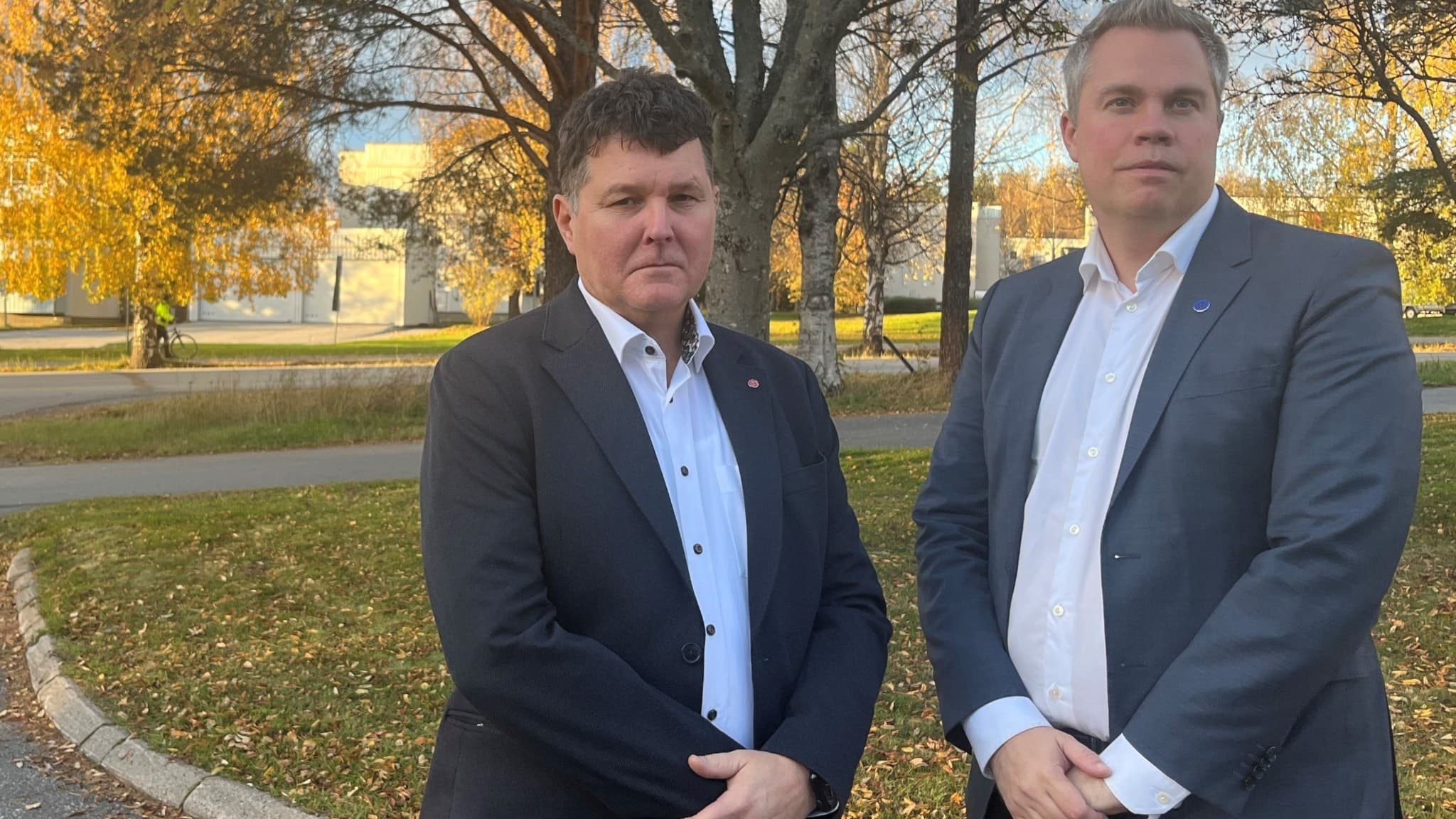 Regionrådet Peter Olofsson (S) och oppositionsrådet Nicklas Sandström (M) står utomhus i höstmiljö med träd och gulnande löv i bakgrunden, båda klädda i kostym.