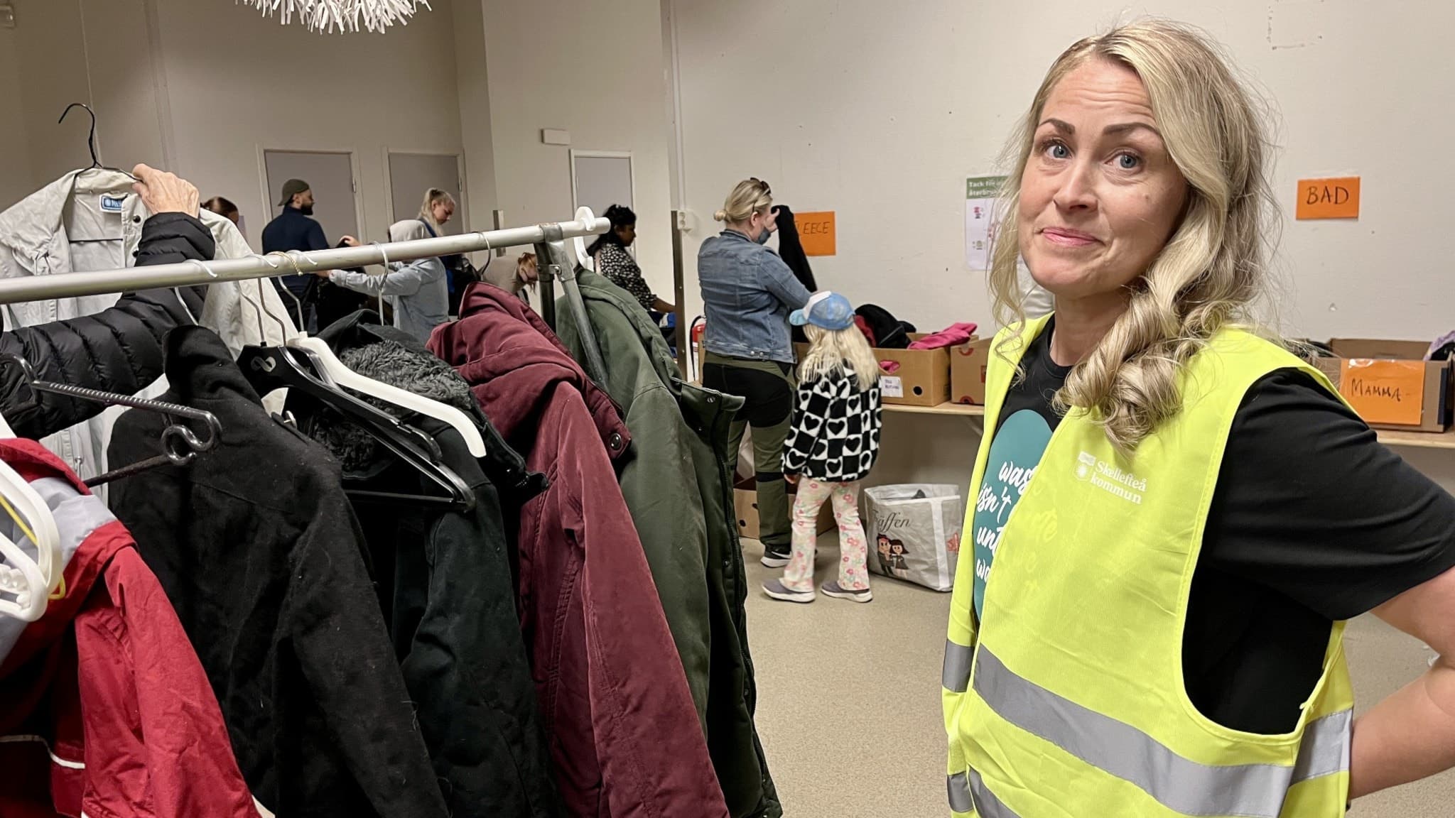 Malin Nyström, klimat- och energirådgivare, står i en lokal med klädställningar fyllda med jackor. I bakgrunden syns personer som sorterar kläder och kartonger i en återbruksinsats.