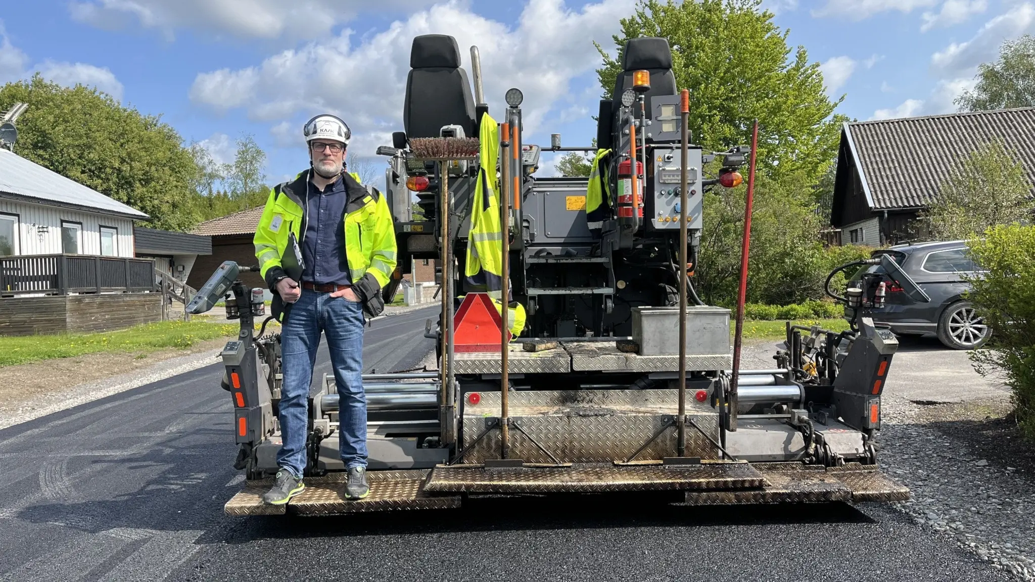 Peter Norberg, projektledare i Härnösand kommun, står på en asfalteringsmaskin med skyddsutrustning. Maskinen är aktiv och omges av nyasfalterad väg, hus och grönska i bakgrunden.