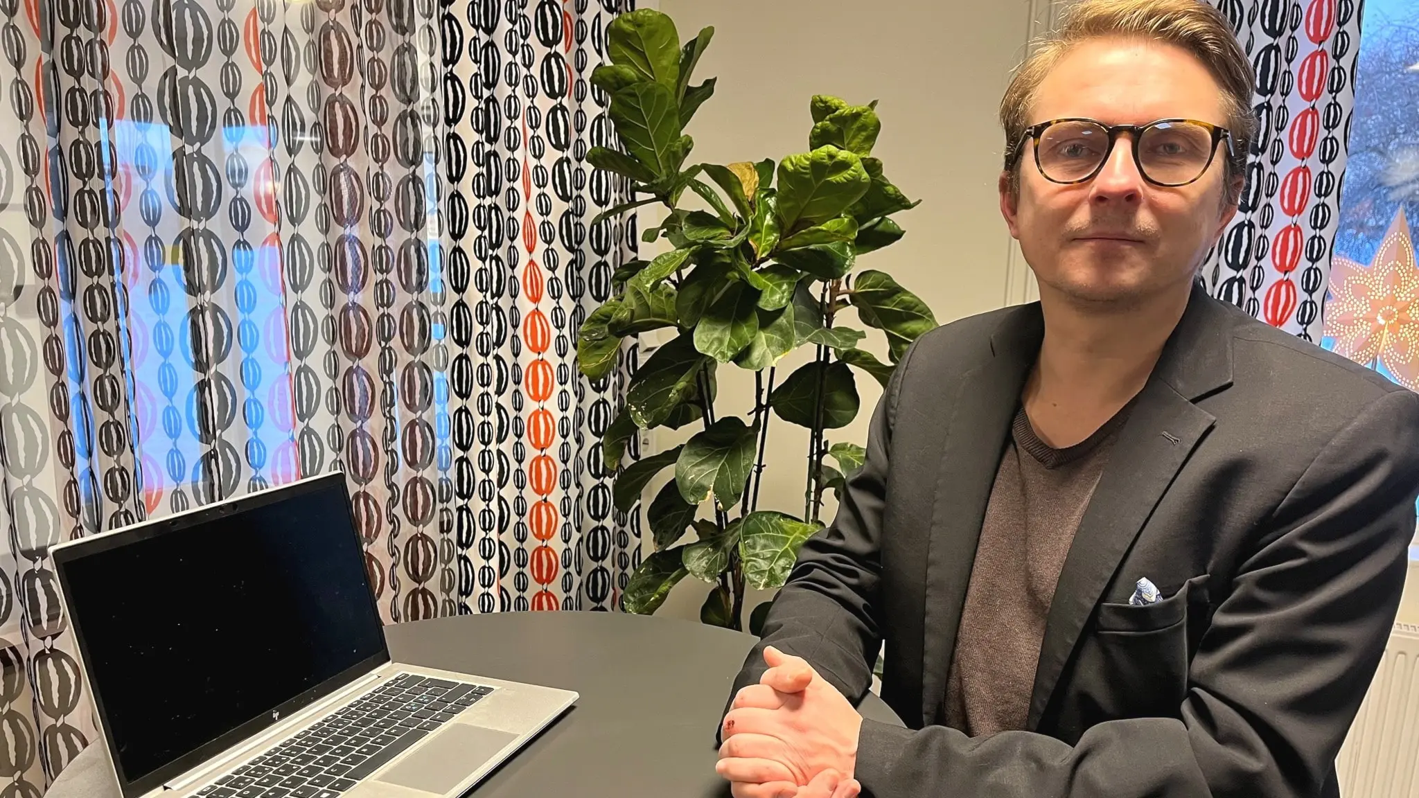 En man klädd i kavaj står vid ett mörkgrått barbord, där en dator står. I bakgrunden syns gardiner och en stor grön växt.