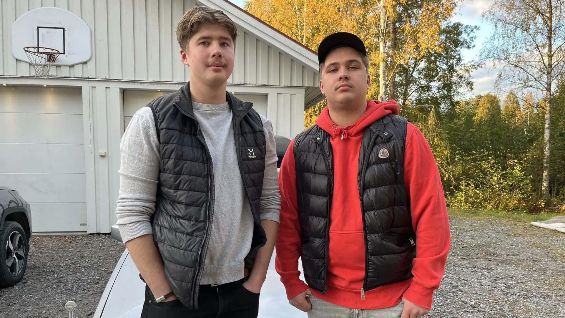 William Olsson Karlsson och Willgot Hirvelä Göransson står utomhus vid en grusplan framför en vit byggnad och en bil, omgivna av höstfärgade träd i Örnsköldsvik.