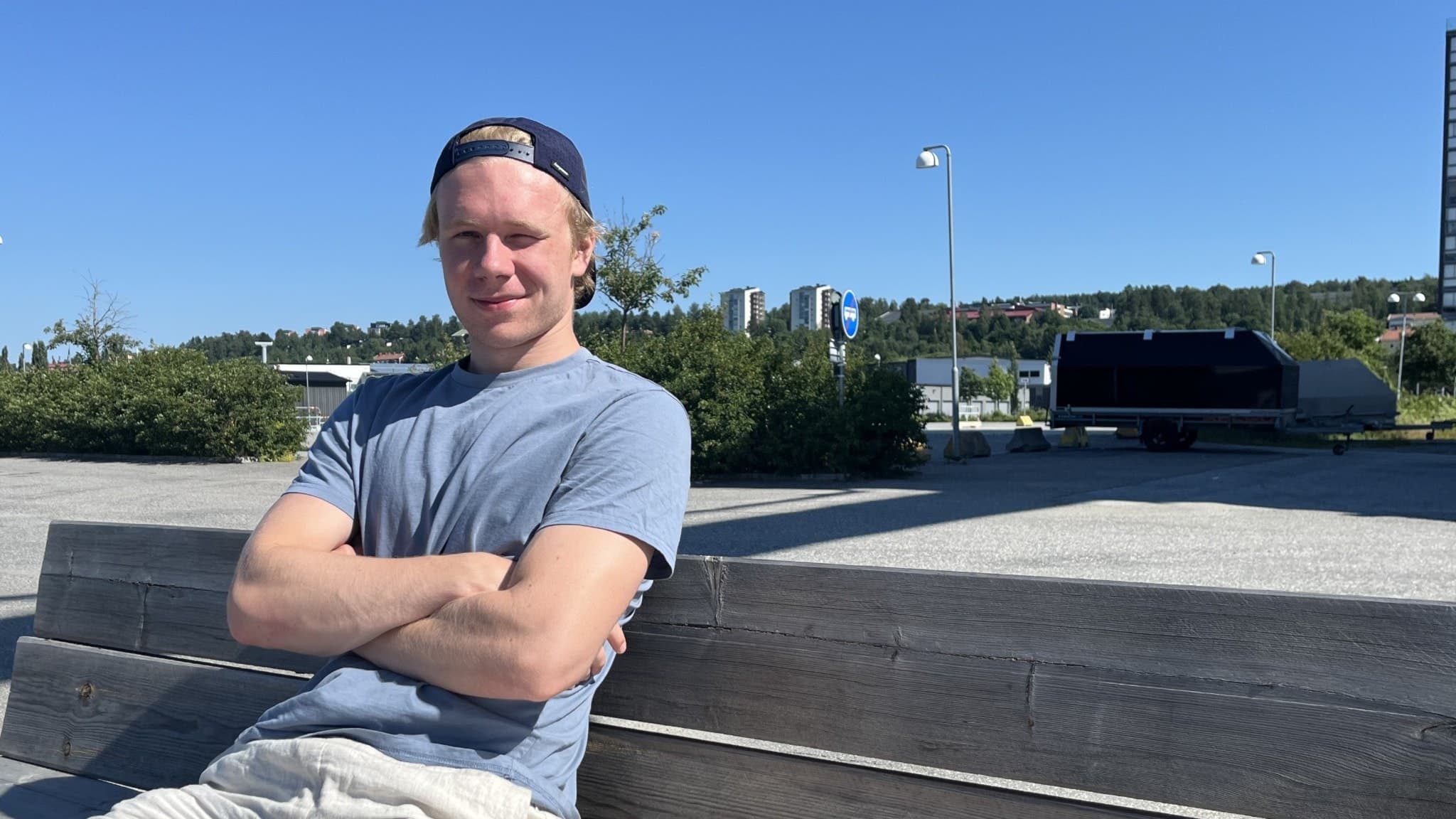 Milton Gästrin från Modo Hockey sitter avslappnat på en träbänk i solskenet, iklädd keps, grå t-shirt och ljusa shorts. En modern byggnad och blå himmel syns i bakgrunden.